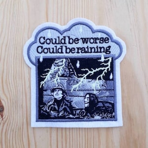 Op de afbeelding: Geborduurde strijkpatch met een grijze wolk en de tekst "Could be worse Could be raining". De wolk heeft regendruppels en een bliksem. De afbeelding onder de wolk toont twee mensen in een donkere scène met een bliksem.