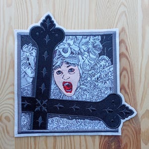 Può includere: Toppa ricamata raffigurante un vampiro e una croce. Il design presenta un'illustrazione dettagliata di un vampiro con labbra e zanne rosse, incorniciata da una croce nera e un bordo decorativo. La toppa è nei toni del grigio e del bianco.