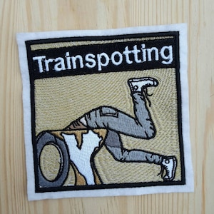 Può includere: Toppa ricamata con sfondo beige e bordo nero. La toppa presenta un contorno bianco di una persona che fa la verticale con le gambe in aria, e il testo "Trainspotting" in lettere bianche su sfondo nero.