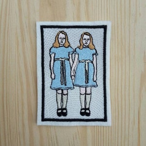 Peut inclure: Patch brodé représentant deux filles en robes bleues avec des cols et des ceintures blancs, debout côte à côte, les mains jointes. Les filles ont les cheveux blonds et sont encadrées d'une bordure noire.