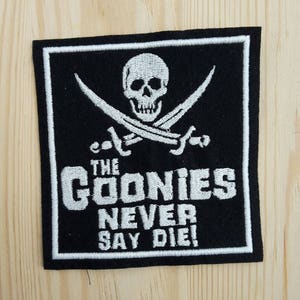 May include: Black and white embroidered patch featuring a skull and crossbones with the text "The Goonies Never Say Die!"