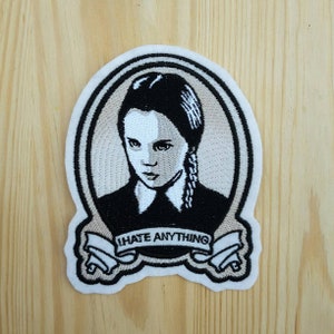 Può includere: Toppa ricamata con il ritratto di una giovane donna con trecce nere, che indossa una camicia nera con colletto. La toppa ha un bordo bianco e uno stendardo con la scritta "I HATE ANYTHING".