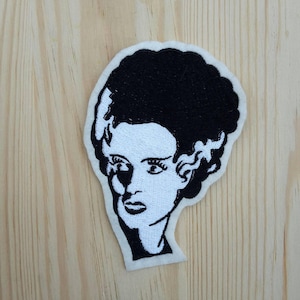 Peut inclure: Patch brodé en noir et blanc représentant le visage d'une femme avec une coiffure rappelant celle de Frankenstein.