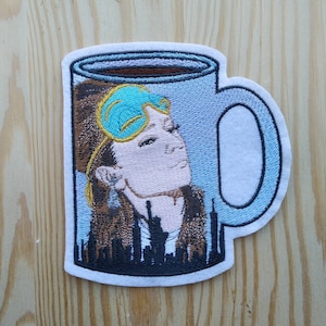 Può includere: Una tazza da caffè ricamata in blu e bianco con il viso di una donna e una maschera per gli occhi blu. La tazza è piena di caffè marrone e ha un bordo bianco.