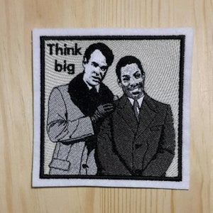 Pode incluir: Patch bordado com dois homens de terno, um com colar de pele, sobre um fundo branco. O texto "Think big" está impresso em preto acima dos homens.