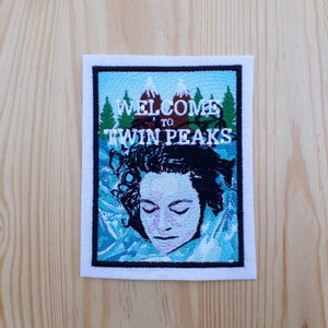 Può includere: Una toppa ricamata con sfondo blu e bordo bianco. La toppa presenta un viso di donna con gli occhi chiusi, circondato da montagne e alberi. Il testo "WELCOME TO TWIN PEAKS" è sopra il viso della donna.