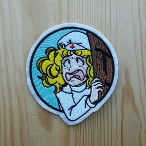 Peut inclure: Patch brodée thermocollante représentant une infirmière de dessin animé aux cheveux blonds, portant une tenue blanche et un chapeau de croix rouge. L'infirmière a une expression effrayée sur son visage et s'accroche à un objet en bois brun.