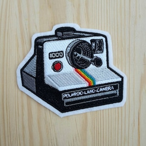 Può includere: Una toppa ricamata di una Polaroid Land Camera in bianco e nero con una striscia arcobaleno sul davanti. La toppa ha un bordo bianco e il testo "POLAROID LAND CAMERA" è ricamato in basso.