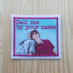 Może przedstawiać: Haftowana naszywka z napisem "Call me by your name" w kolorze różowym. Obraz przedstawia dwie postacie patrzące w górę, na jasnoniebieskim tle, z różową obwódką i białym konturem.