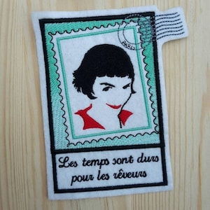 Könnte beinhalten: Gestickter Patch mit grünem Rand und weißem Hintergrund. Der Patch zeigt eine Schwarzweißillustration einer Frau mit rotem Shirt. Der Text "Les temps sont durs pour les rêveurs" ist in Schwarz unter der Illustration geschrieben.