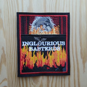 May include: Black embroidered patch with the text "INGLOURIOUS BASTERDS" in white. The patch features a stylised image of a person's head engulfed in flames.