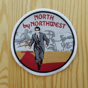May include: A white circular embroidered patch with a red, white and gold design. The patch features a man in a suit running on a red carpet with a plane in the background. The text "NORTH by NORTHWEST" is embroidered in red on the patch.