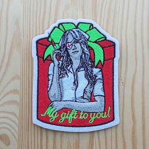 Può includere: Toppa ricamata raffigurante una donna con cappello da infermiera e benda sull'occhio, che tiene una siringa. L'immagine è ambientata in un cofanetto rosso con un fiocco verde, e il testo "My gift to you!" è scritto in verde.