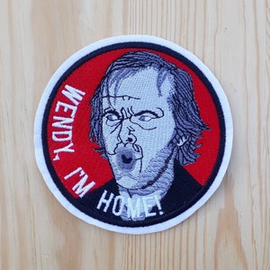 Può includere: Una toppa ricamata bianca e rossa con un'immagine in bianco e nero del personaggio di Jack Nicholson del film "Shining" con il testo "Wendy, I'm Home!"