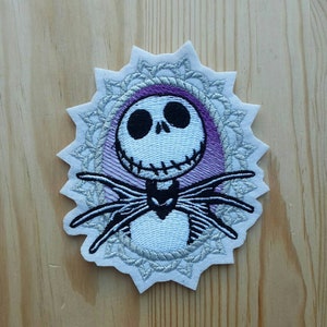 Può includere: Toppa ricamata da stirare con un Jack Skellington bianco e nero di The Nightmare Before Christmas. La toppa è circondata da un design a mandala argentato e bianco con sfondo viola.
