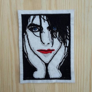 Op de afbeelding: Zwart-wit geborduurde patch van een persoon met lang zwart haar, blauwe ogen en rode lippen. De persoon kijkt de kijker aan met zijn handen op zijn gezicht.
