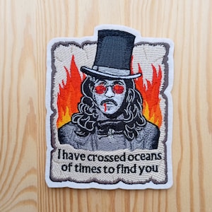 Può includere: Toppa ricamata con un vampiro con un cappello a cilindro e il naso sanguinante. Il vampiro è circondato da fiamme e dal testo "Ho attraversato oceani di volte per trovarti".