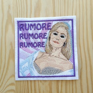 Op de afbeelding: Een geborduurde strijkpatch met een vrouw met blond haar en de tekst "Rumore Rumore Rumore" in paarse letters op een roze en paarse achtergrond.