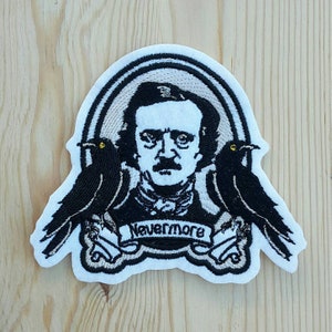Peut inclure: Patch brodé représentant un portrait d'Edgar Allan Poe avec deux corbeaux noirs de chaque côté. Le patch a un fond blanc et le texte "Nevermore" est écrit sur une bannière sous le portrait de Poe.