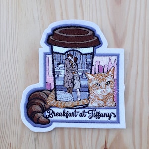 Op de afbeelding: Geborduurde patch met een koffiebeker met een scène van een stel dat voor een taxi kust, een kat en de tekst "Breakfast at Tiffany's".