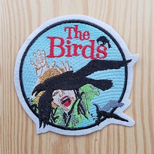 Può includere: Una toppa rotonda ricamata bianca su sfondo blu. La toppa presenta un uccello nero che attacca una persona con il testo "The Birds" in lettere rosse.