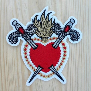 Può includere: Toppa ricamata con un cuore rosso trafitto da due spade argentate, circondata da un bordo di perline dorate e bianche. Una fiamma dorata si trova sopra il cuore.