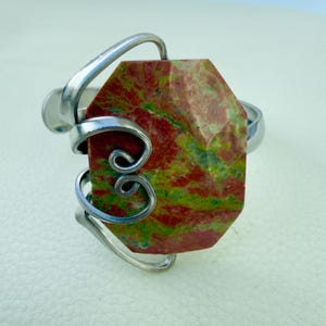 May include: A silver cuff bracelet featuring a large, faceted unakite stone. The stone displays a mix of red and green hues. The bracelet has a decorative, swirling silver design around the stone, adding a unique touch to the jewelry.