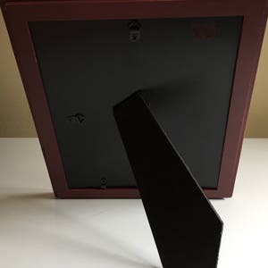 May include: A dark red picture frame with a black mount and a black easel stand. The frame has two hanging hooks on the back.