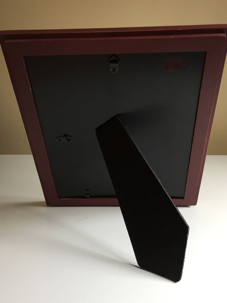 May include: A dark red picture frame with a black mount and a black easel stand. The frame has two hanging hooks on the back.