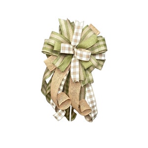 May include: A decorative bow featuring a mix of green, white, and tan ribbons. The bow has a layered design with gingham and striped patterns, and burlap-style ribbons. The bow is suitable for various decorative purposes.
