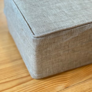 May include: A close-up of a gray fabric ottoman with a stitched seam around the top edge.