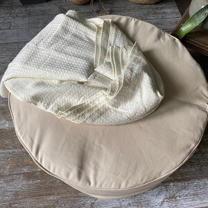 May include: A round, beige, fabric cushion with a white, textured blanket on top. The cushion is on a wooden floor.