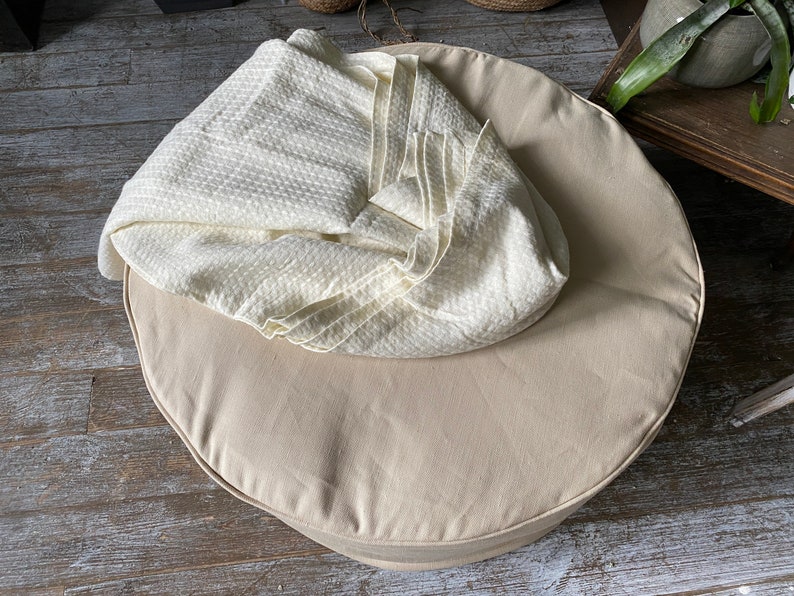 May include: A round, beige, fabric cushion with a white, textured blanket on top. The cushion is on a wooden floor.