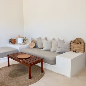 May include: A light gray couch with a wooden frame and a light gray cushion. The couch is in a white room with a white wall and a white floor. There is a wooden coffee table in front of the couch with a brown tray on top. There is a rug on the floor in front of the couch.