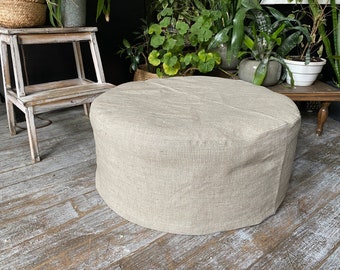 May include: A round, light beige linen ottoman with a smooth, textured surface. The ottoman is sitting on a light wood floor.