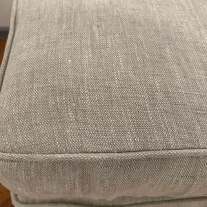 May include: Close-up of a light grey linen fabric with a subtle texture. The fabric has a stitched edge.