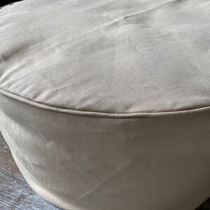 May include: A round, beige fabric ottoman with a smooth, textured surface. The ottoman has a simple, classic design.