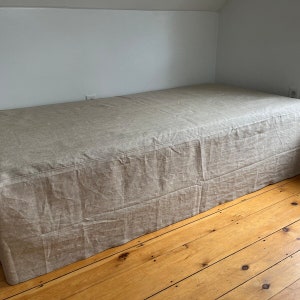 May include: A beige linen bed skirt with a simple design. The skirt is draped over a wooden floor.