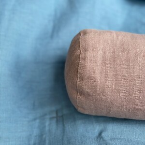 以下が含まれることがあります： リネン生地のカバーが付いた薄い茶色の円筒形の枕。枕は青い布の表面に置かれています。