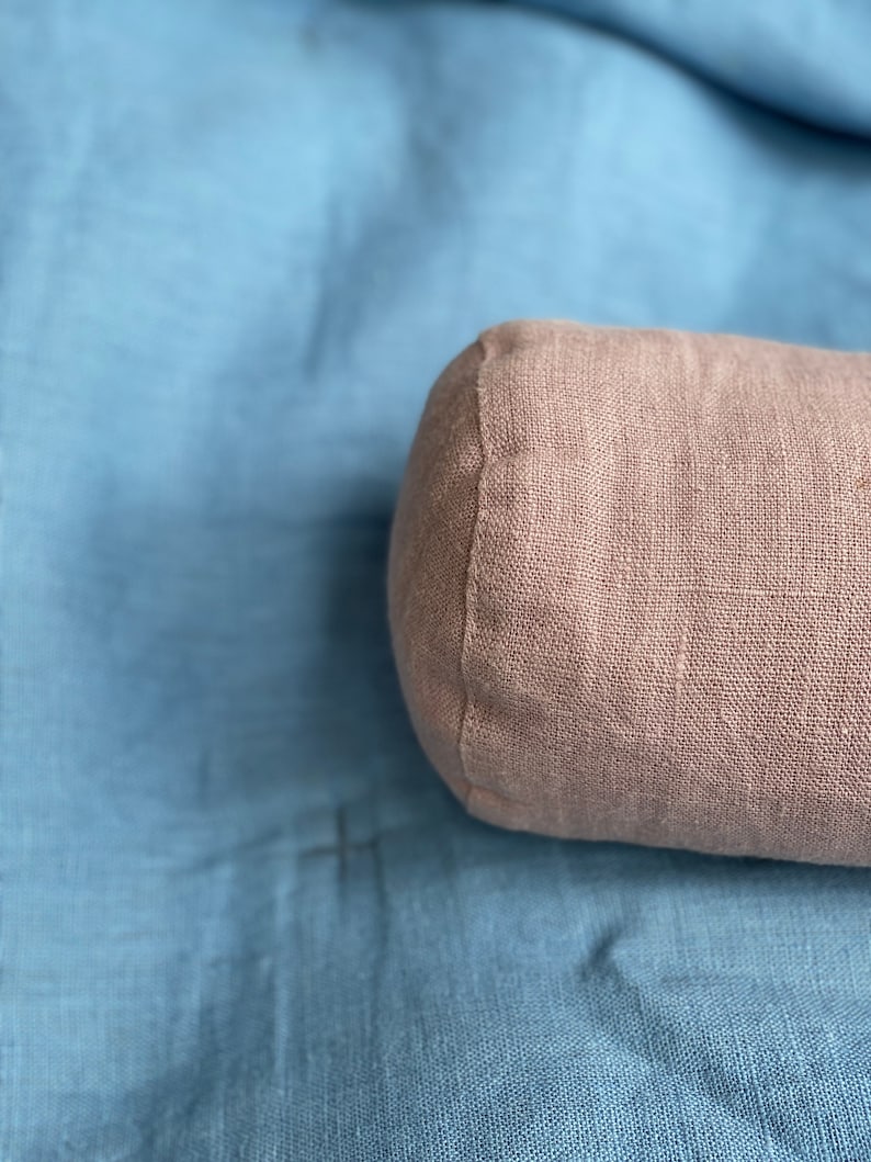 以下が含まれることがあります： リネン生地のカバーが付いた薄い茶色の円筒形の枕。枕は青い布の表面に置かれています。