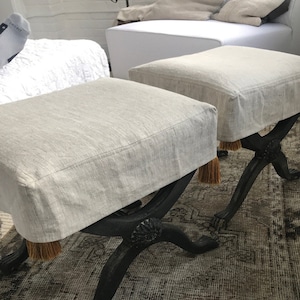 May include: Two grey linen-covered ottomans with black wooden legs and tassel fringe. The ottomans are on a patterned rug.