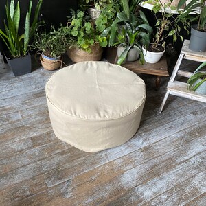 May include: A round, beige, fabric-covered ottoman sits on a wooden floor. The ottoman is in focus, while the background is blurred and features plants in pots.