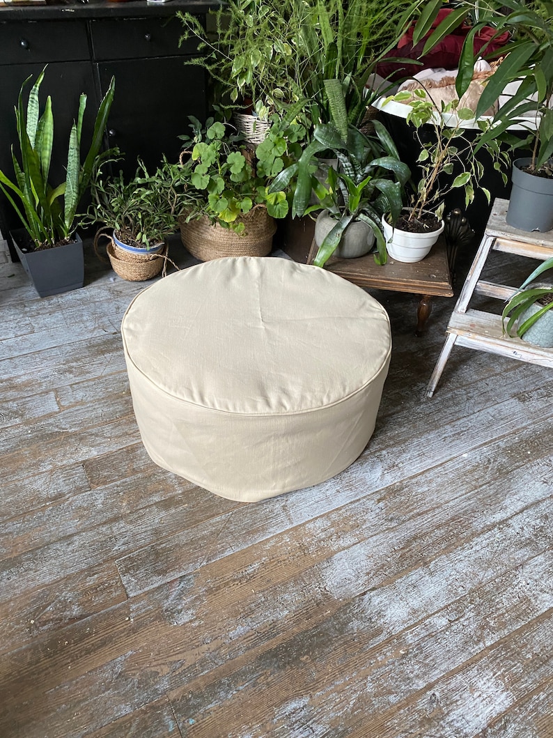 May include: A round, beige, fabric-covered ottoman sits on a wooden floor. The ottoman is in focus, while the background is blurred and features plants in pots.