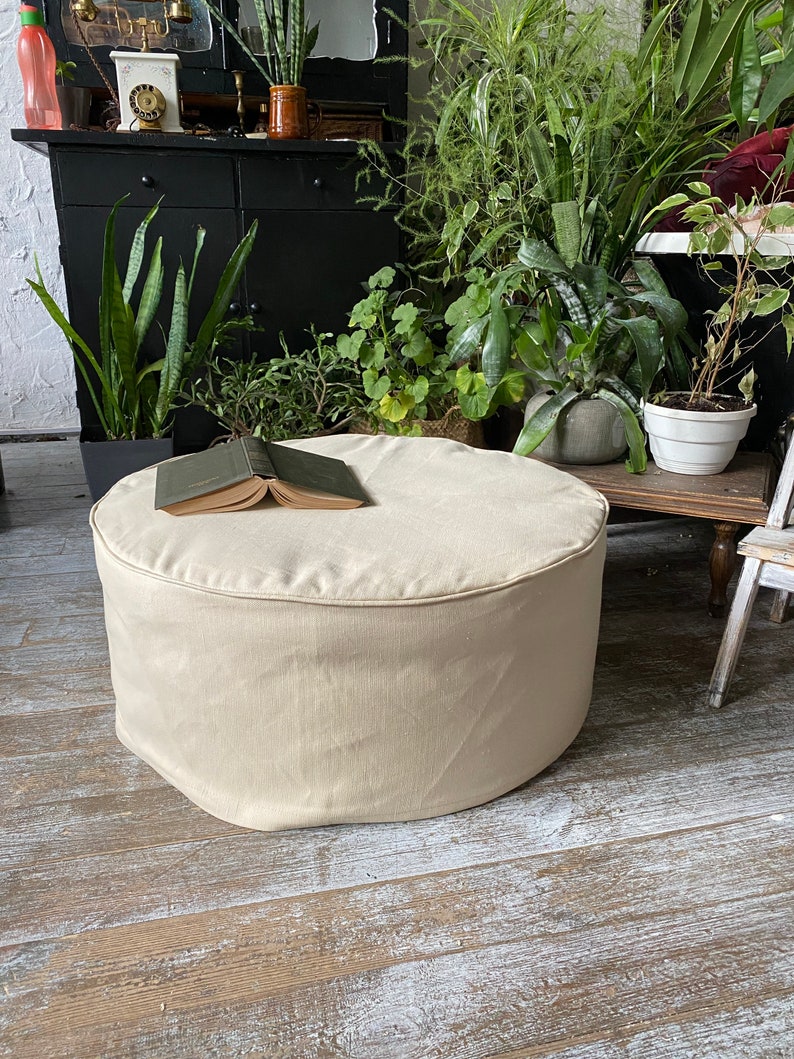 May include: A cream-colored round ottoman with a book on top. The ottoman is sitting on a wooden floor.