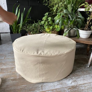 May include: A round, beige, fabric-covered ottoman with a smooth surface. The ottoman is sitting on a light wood floor.