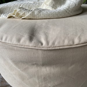 May include: A round, beige ottoman with a white throw blanket draped over it. The ottoman is made of a soft, textured fabric.