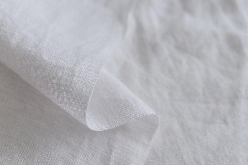 May include: Close-up of a white linen fabric with a soft, textured surface. The fabric is folded and draped, creating a smooth, flowing curve.
