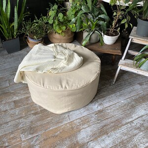 May include: A beige round ottoman with a white throw blanket on top. The ottoman is sitting on a wooden floor with white paint splatters.