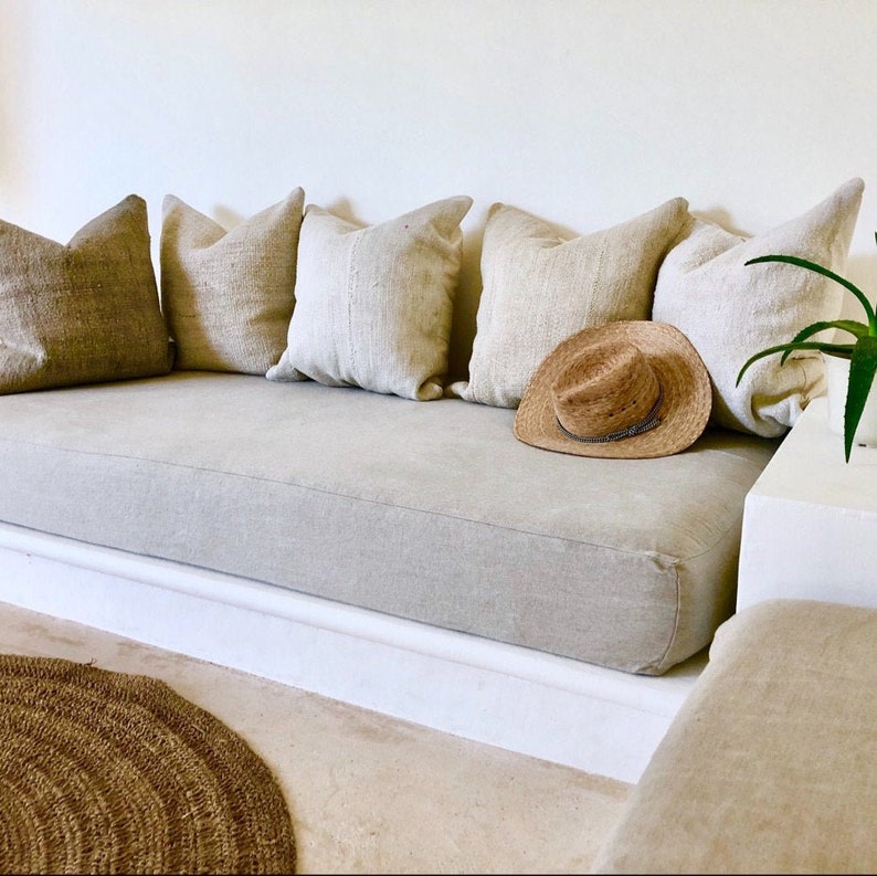 May include: A light beige linen sofa with a white base and a straw hat on the seat. The sofa is covered in several beige pillows.