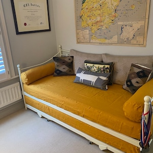 May include: A white metal daybed with a mustard yellow linen mattress and pillows. The daybed has a white metal frame and a white metal headboard. There are several pillows on the daybed, including a gray pillow with a white fox design, a brown and black patterned pillow, and a white pillow with a floral design.